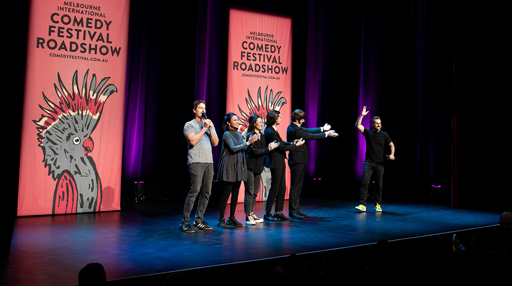 Image of five comedians on stage with two large banners behind them with the Melbourne International Comedy Festival Roadshow logo on them